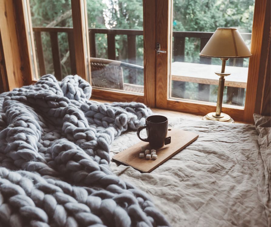 Early light shining into a cozy bedroom. A heavy blanket and hot chocolate on a bed facing windows.