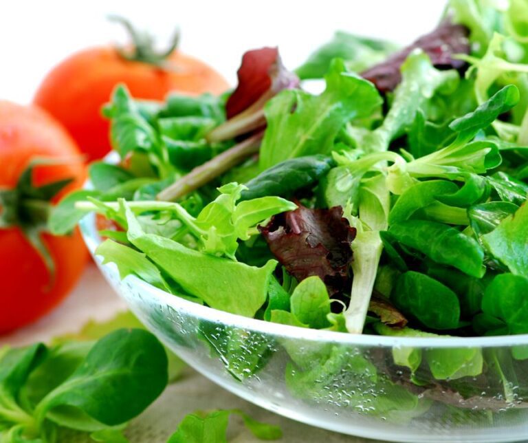 A clear bowl of fresh multi colored baby greens next to two red ripe tomatoes.