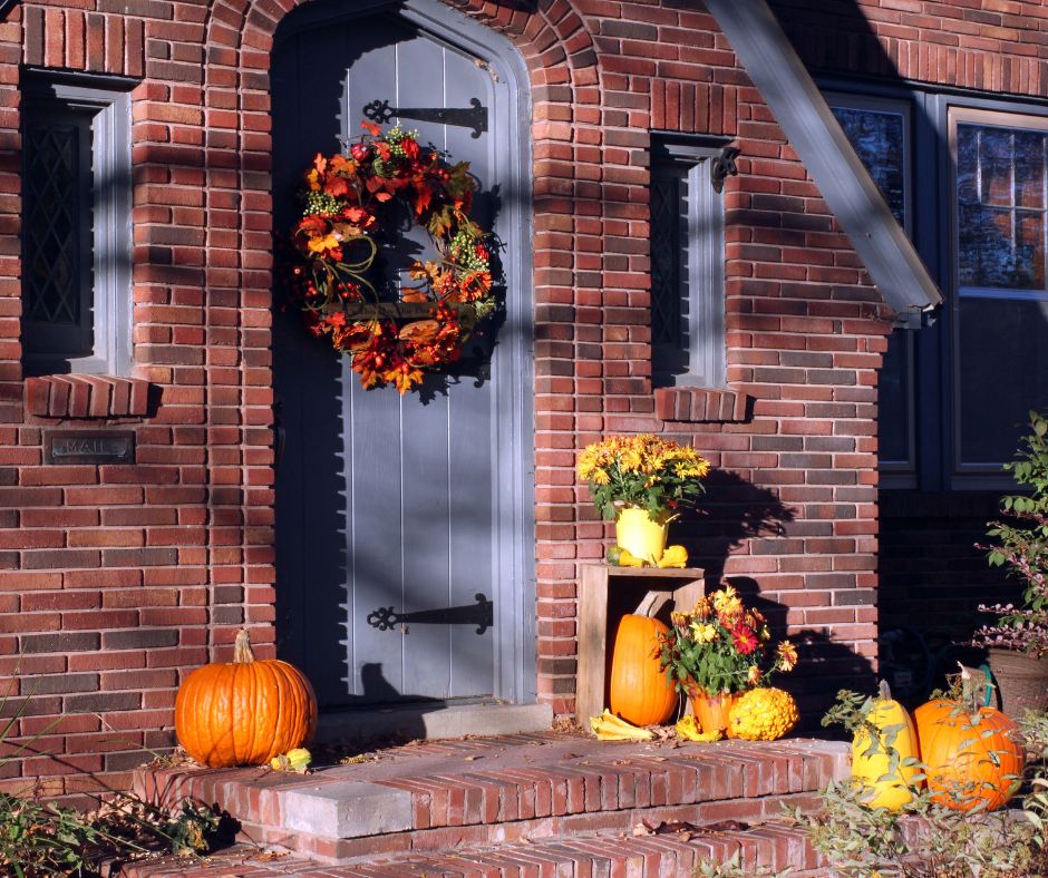 A large wreath, pumpkins and mums decorating the front porch of a cottage.