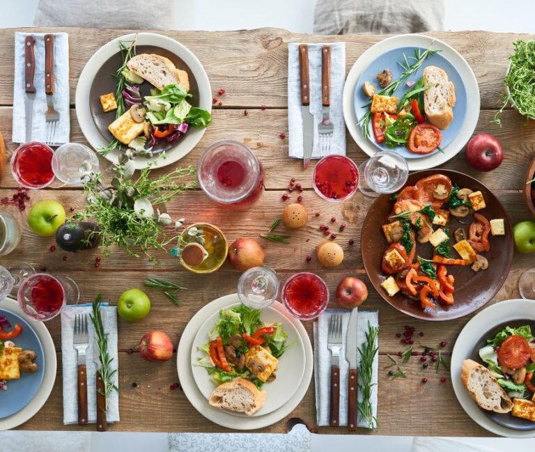 A wooden table set with a fall dinner theme. Including roasted chicken, veggies, and red drinks,