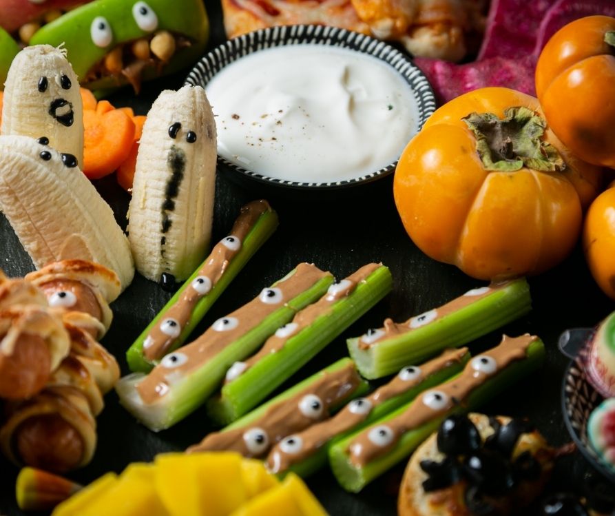 A white herbal dip in a black cup surround by Halloween theme decorated veggies and fruit.