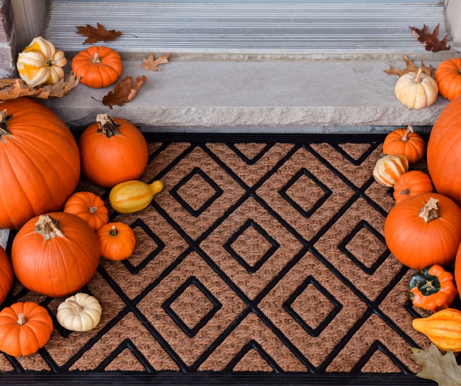 A natural fiber doormat with black diamond pattern edged by an assortment of small gourds and pumpkins.