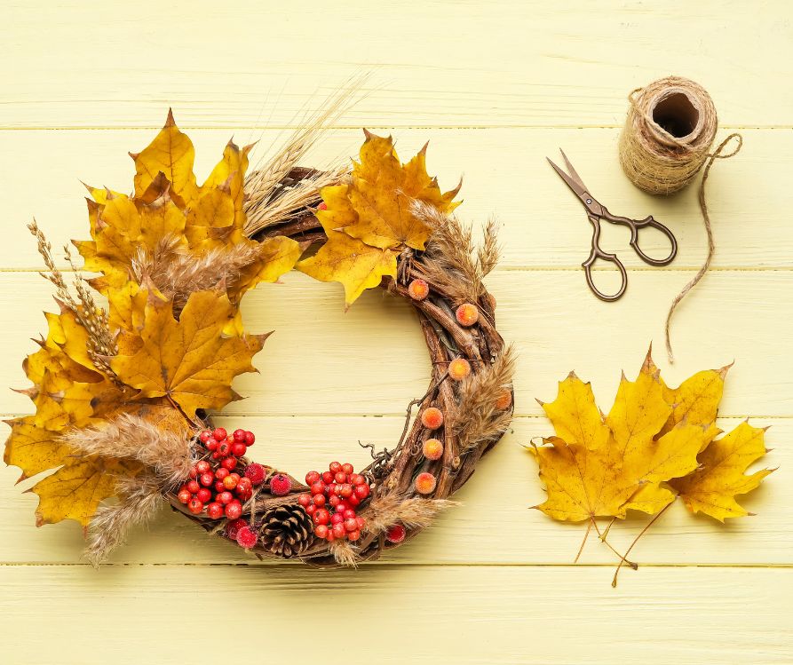 A partially finished autumn wreath on a wood table next to twine, scissors, and yellow preserved leaves.