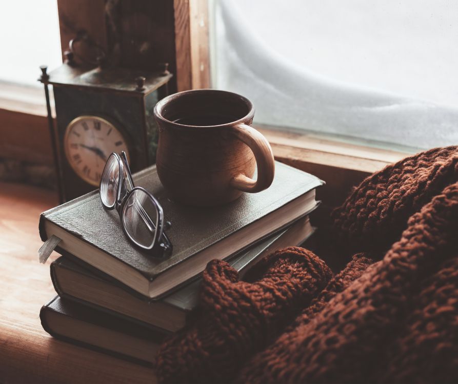 Coffee, eyeglasses, maroon blanket on a deep window seal next to a winterized window.