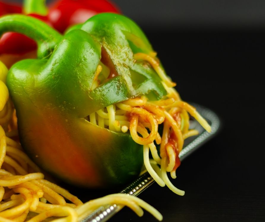 A green bell pepper with a spaghetti coming out of the face cut into its side.