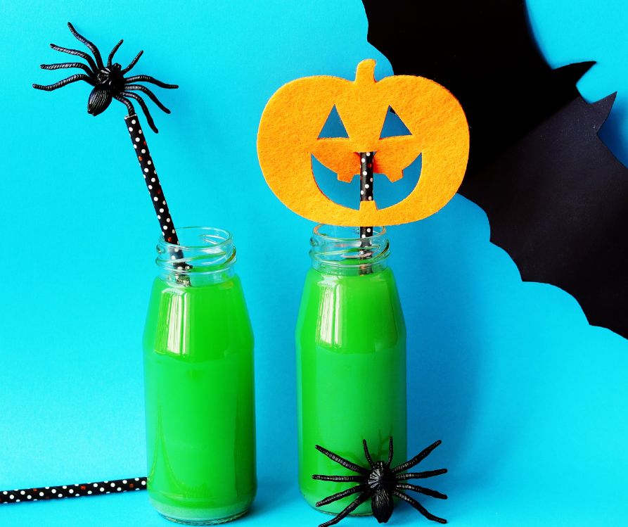Bottles filled with green smoothie on blue background surround by toy spiders, a bat, and an orange jack o lantern cutout.
