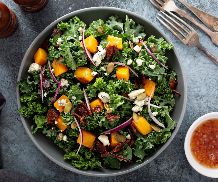 Dark green kale salad topped with bacon and roasted butternut squash in a dark grey plate.