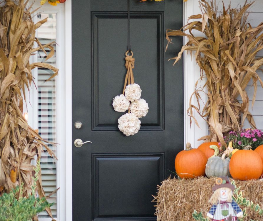 An entryway framed by haybales, dried corn stalks, pumpkins and figurines. A black door is adorned by a door hanger of dried white flowers.
