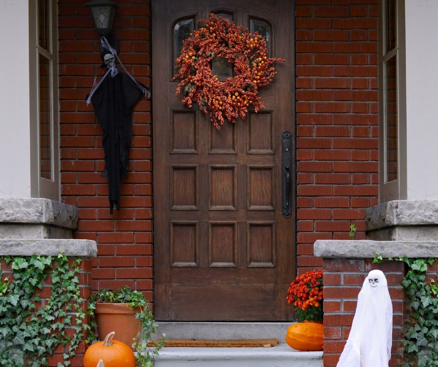 A front porch decorated with orange pumpkins, red potted flowers, a white ghost, a hanging witch and a red berry wreath.