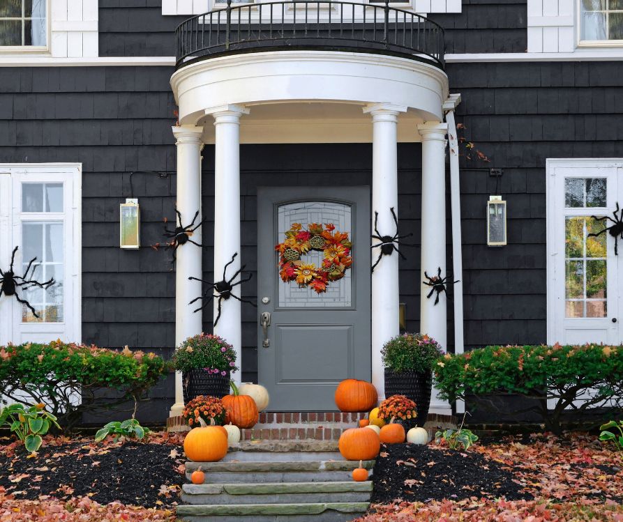 Large black spider decorations place on the exterior of a home. Red fall wreath on door and orange pumpkins on steps.