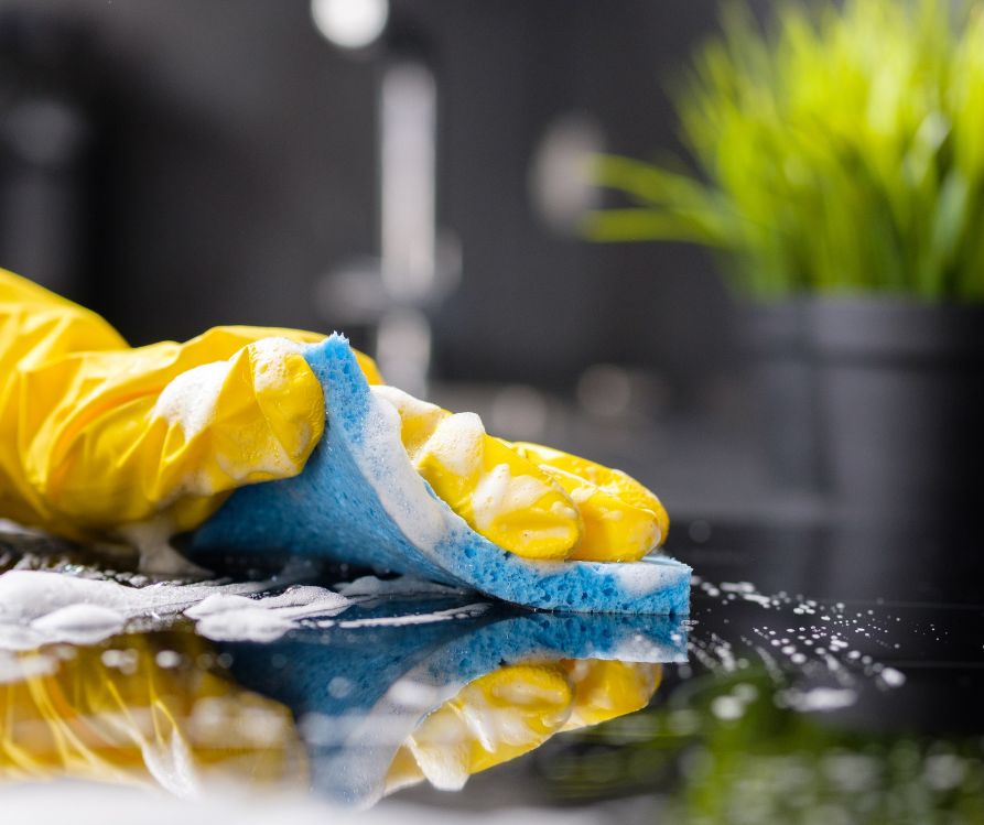 A person wearing yellow gloves wiping down a dark counter top with a blue sponge.