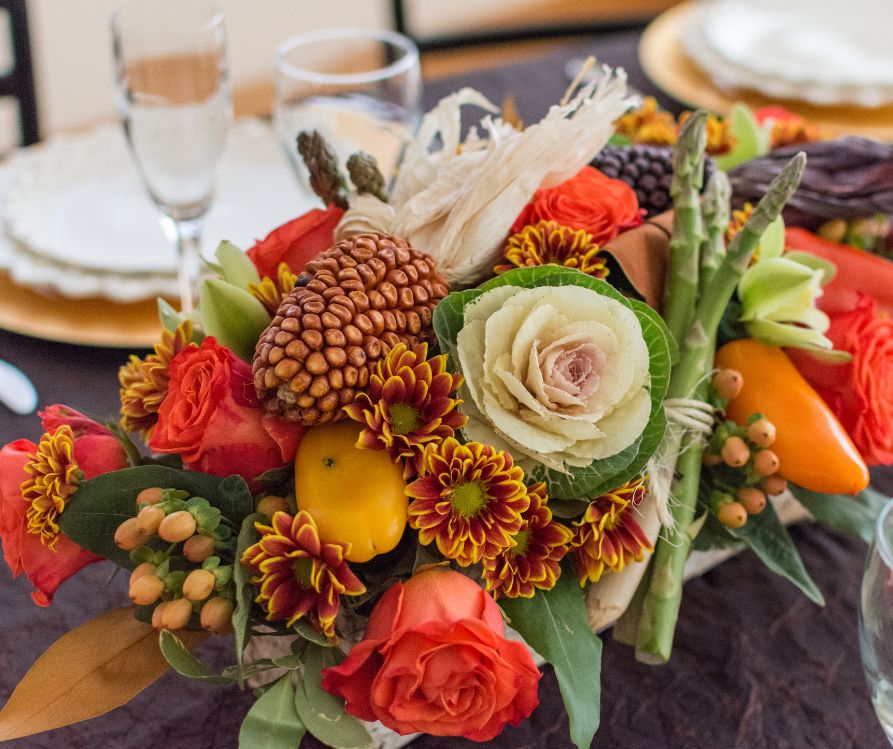 Orange flowers, pinecones and small fall veggies arrangement at the center of a set dinning table.