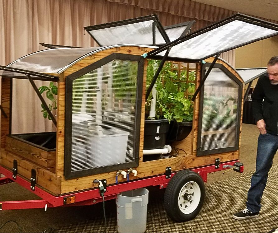 A man checking out an aquaponics garden system built on a trailer with a mini greenhouse on top.
