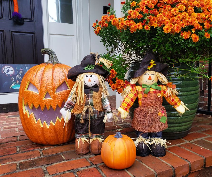 Two small scarecrow dolls next to a large orange jack-o-lantern and small pumpkin.