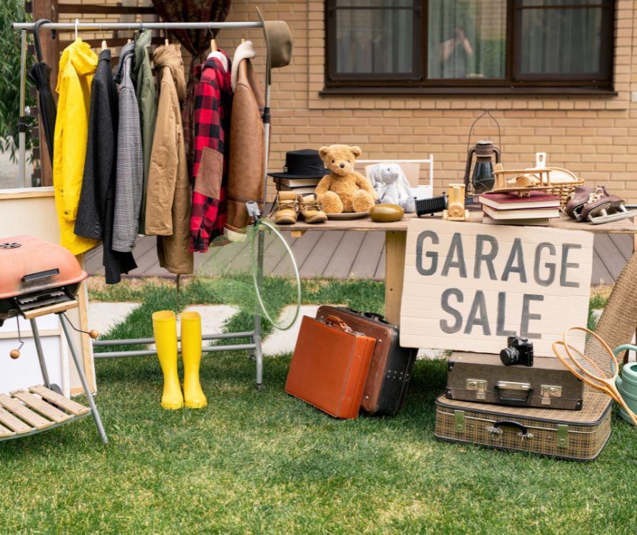 Household items like cloths, shoes, and sports gear on a lawn next to a sign reading "Garage Sale."
