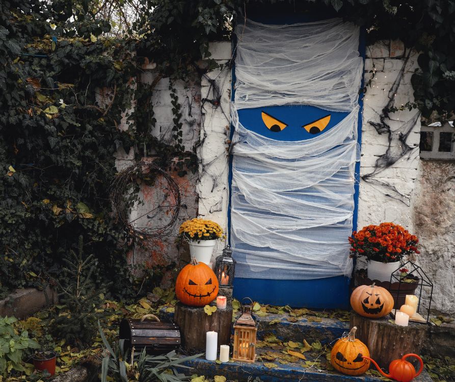 A blue door covered in white cloth to look like a mummy with large yellow eyes. Orange jack-o-lanterns decorate the stairs in front of it.