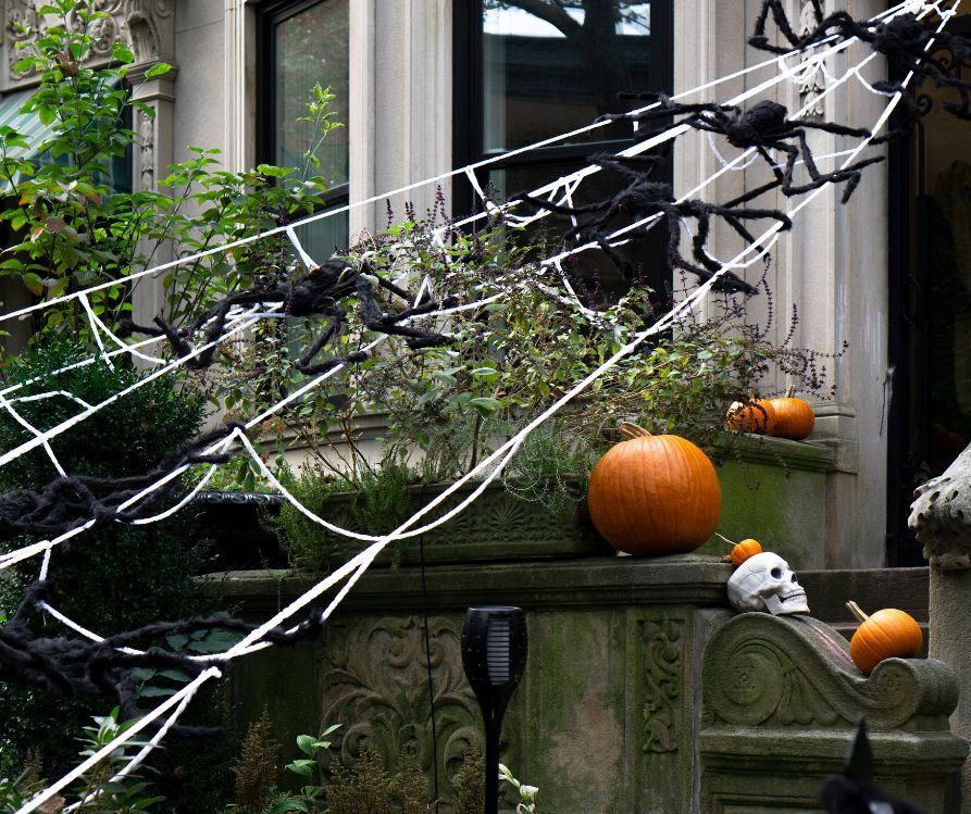 A large spider web made out of white yarn and black spiders decorate a front door along with orange pumpkins and white skulls.
