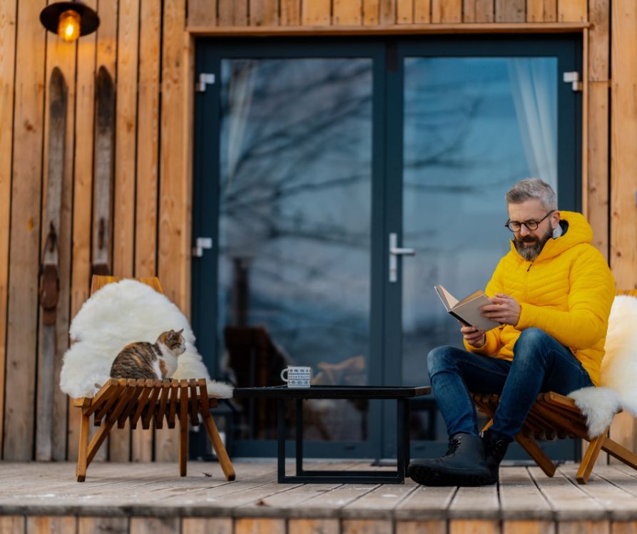 A man in a yellow jacket reading a book reading a book with a cat on a patio. Both are sitting in a wooden chairs.