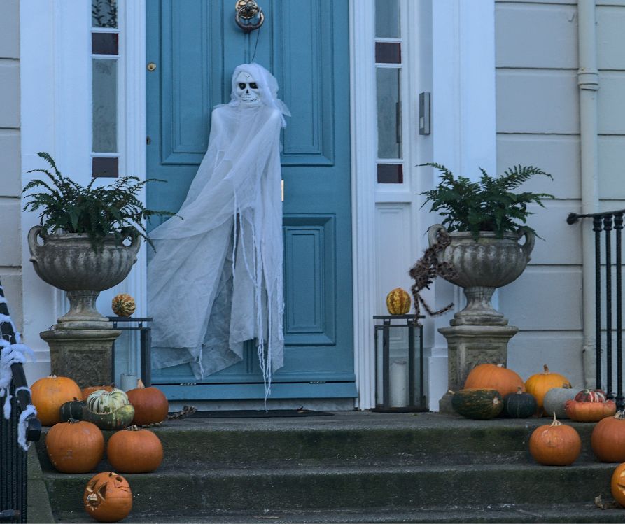 A white ghost door hanger on a light blue front door next to potted ferns and orange pumpkins on the stairs.