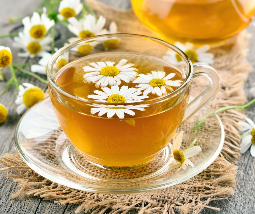 A cup of tea in a clear cup with fresh chamomile flowers floating on the surface.