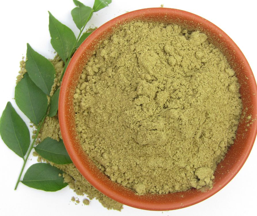 Powdered curry leaves in a bowl on white background next to fresh curry leaves.