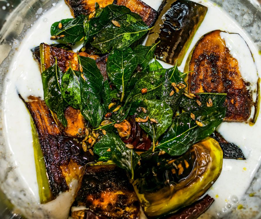 An Indian dish made with brinjal curd curry in oil fried with curry leaves, red chili, cumin, mustard seeds, and Fenugreek seed on a white plate.