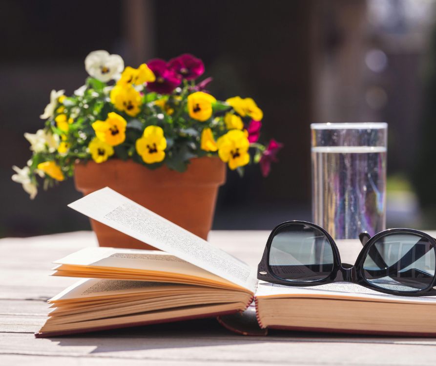 A pot of flowering pansies on a patio table next to a book, sunglasses, and a glass of water.
