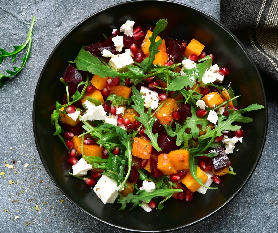 A mixed salad featuring arugula, pomegranate seeds, sweet potatoes, and goat cheese in a black bowl.
