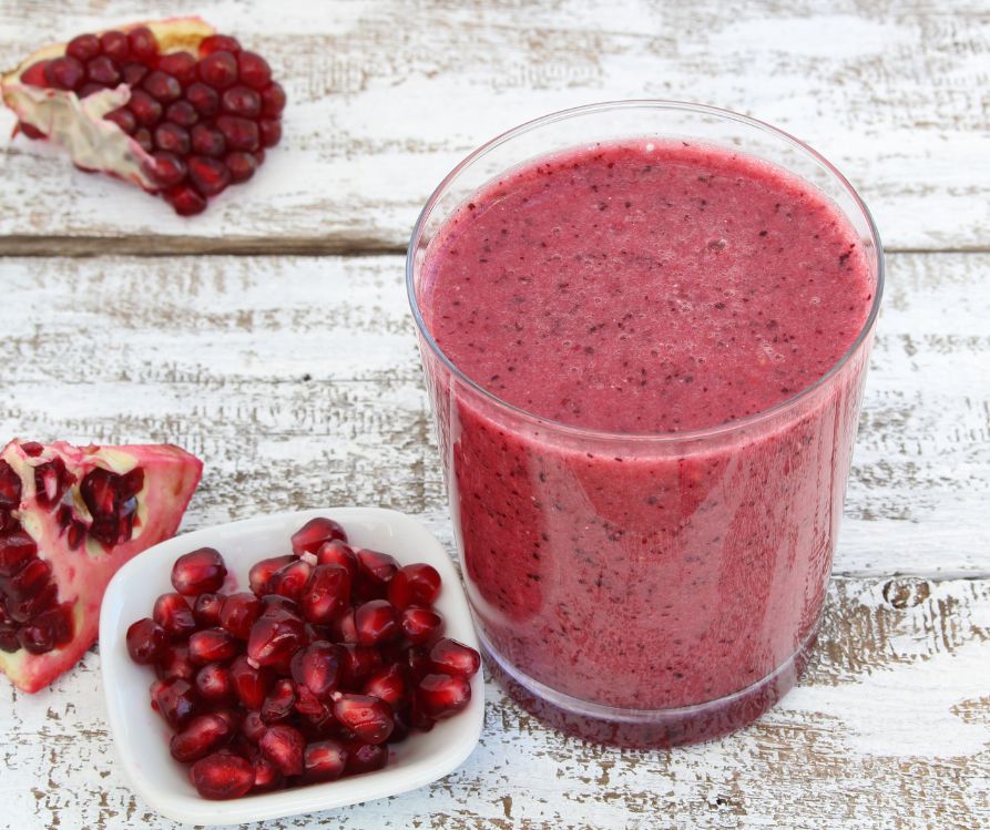 A pink smoothie in a clear glass next to pomegranate seeds.