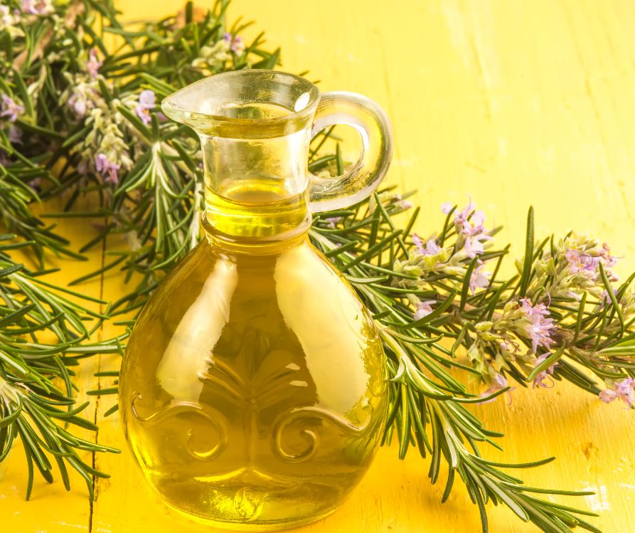 A glass container of olive oil next to fresh rosemary on a yellow table.