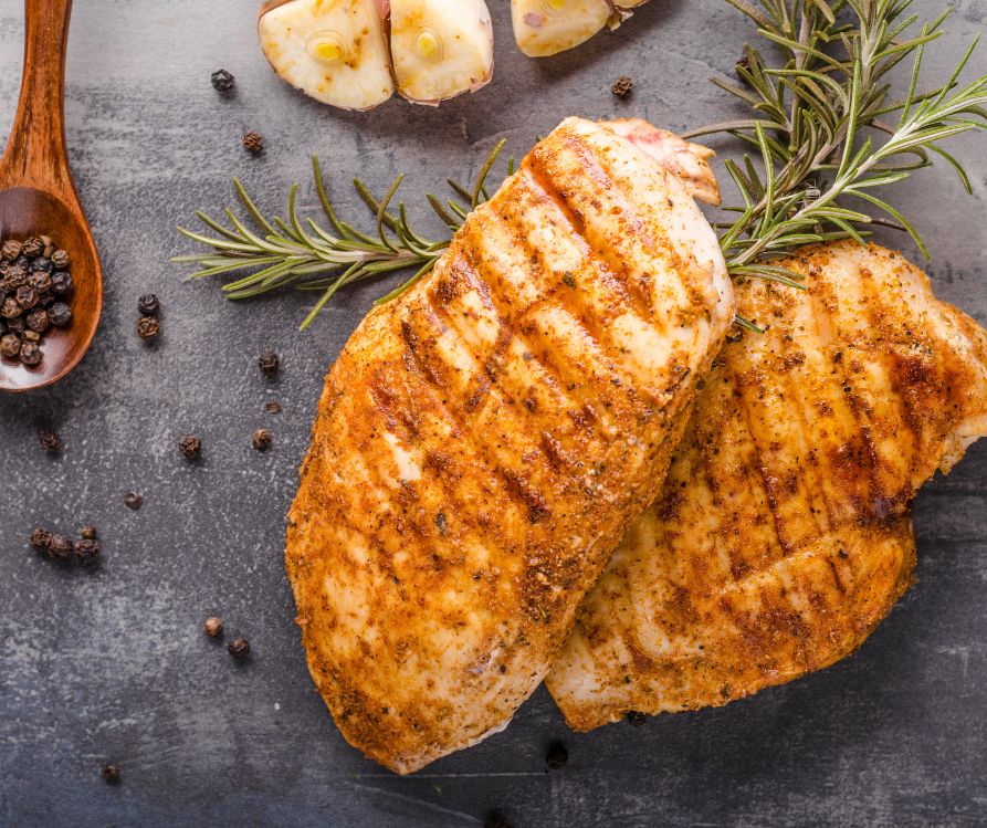 Cooked chicken breast on a black countertop with black peppercorns, rosemary sprigs, and garlic.