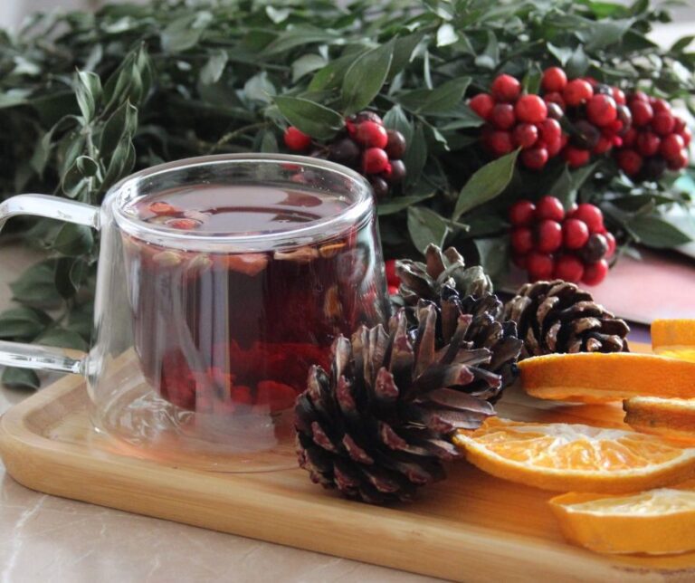 Winter tea in a clear mug next to holy, pinecones and fresh orange slices.
