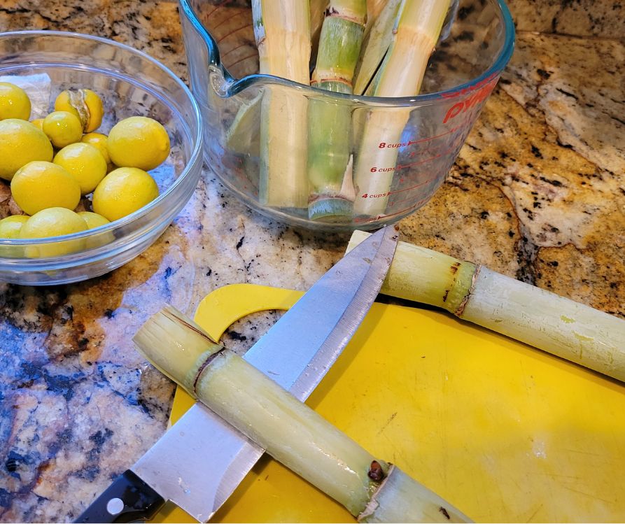 A knife cutting a sugarcane in half on a yellow cutting board. Plus a bowl of small limes and a container of cut sugarcane on a kitchen counter.