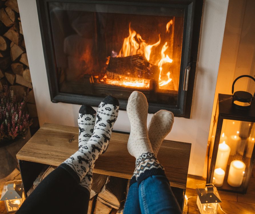 Two people warming their feet next to a fireplace.