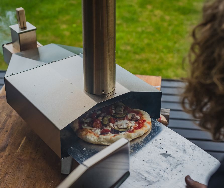 A person loading homemade pizza into an backyard pizza oven.