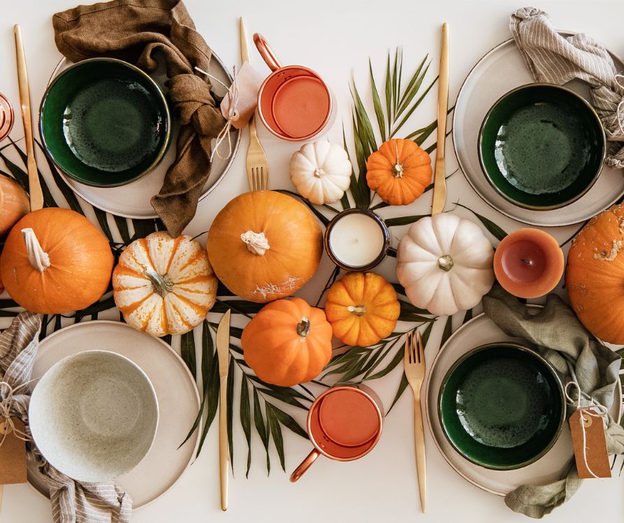 A natural table setting consisting of neutral tone tableware, centerpiece of plant fronds small pumpkins and candles.