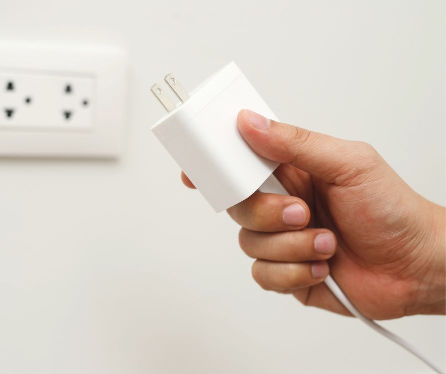 A hand hold a while device charger next to a wall outlet.