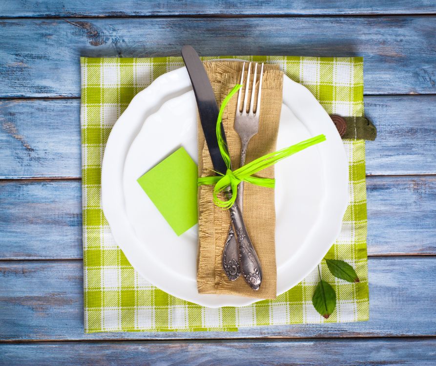 A lime green checkered fabric place mat, white plate and natural napkin wrapped around  silverware on a blue wooden table.