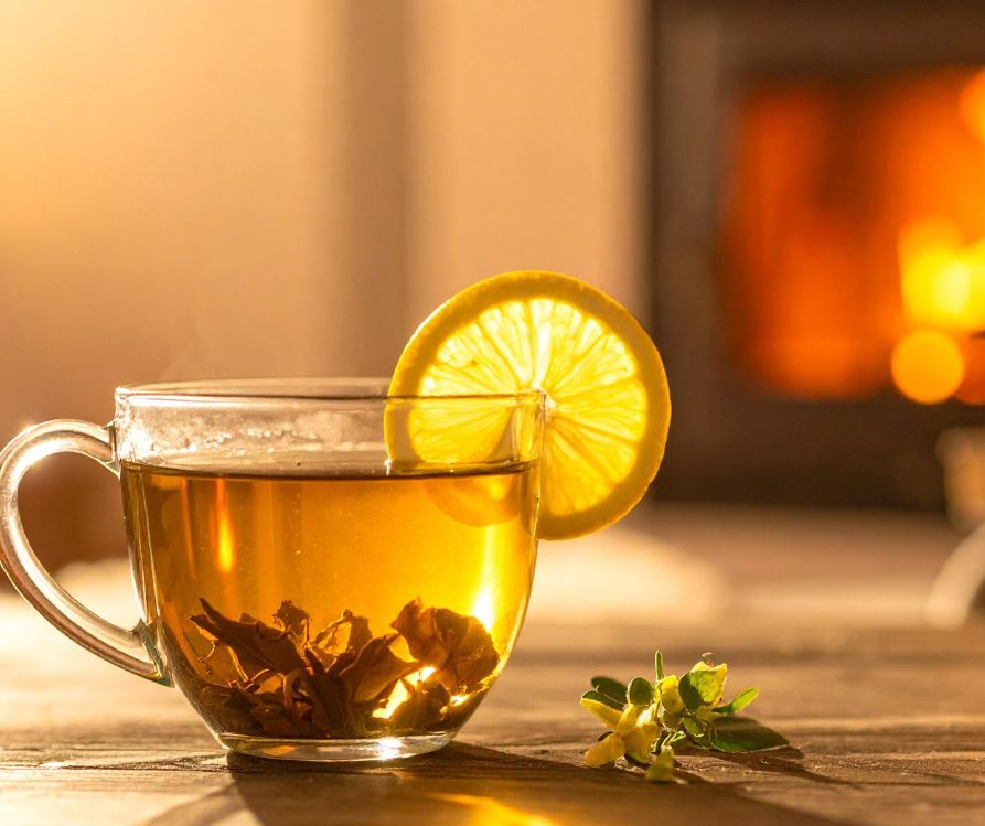 A clear cup of tea with lemon on a table next to a roaring fireplace.