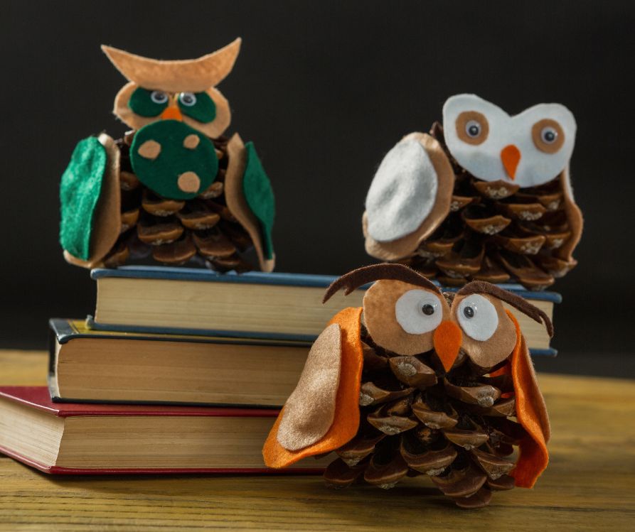 Small owls made of pinecones and felt posed next to a pile of books.