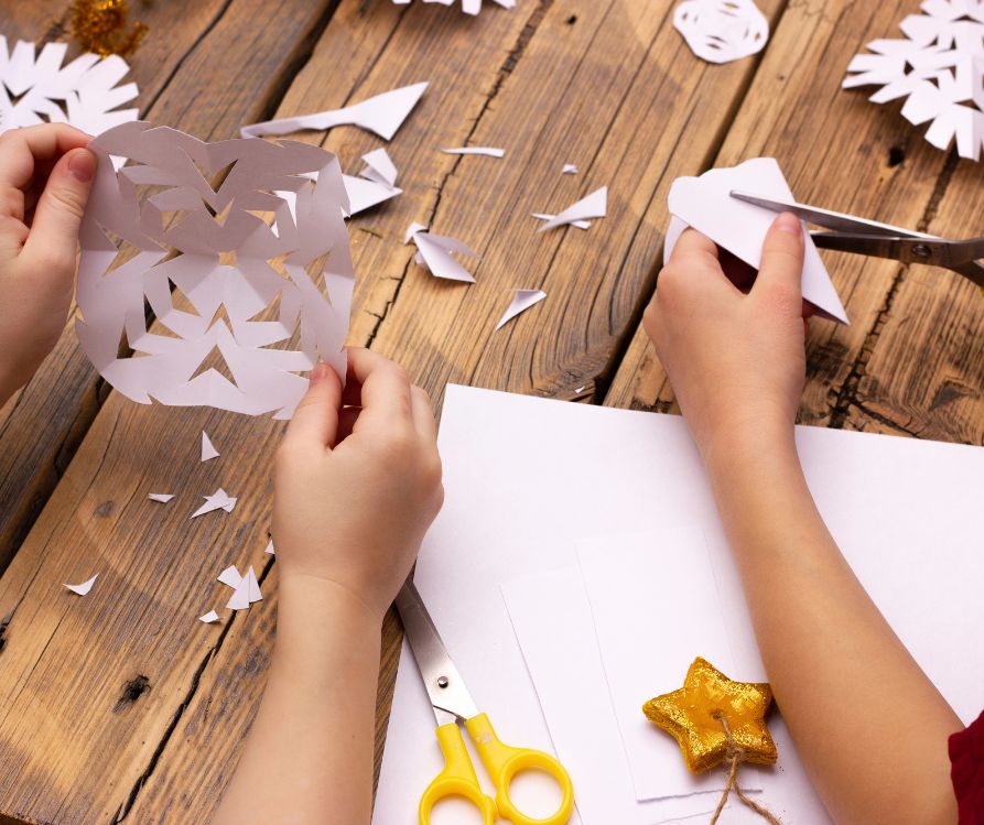 Kids making snowflakes out of white paper at a wooden table.