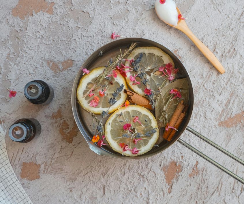 A pot with citrus slices, spring flowers, and herbs next to two essential oil bottles.