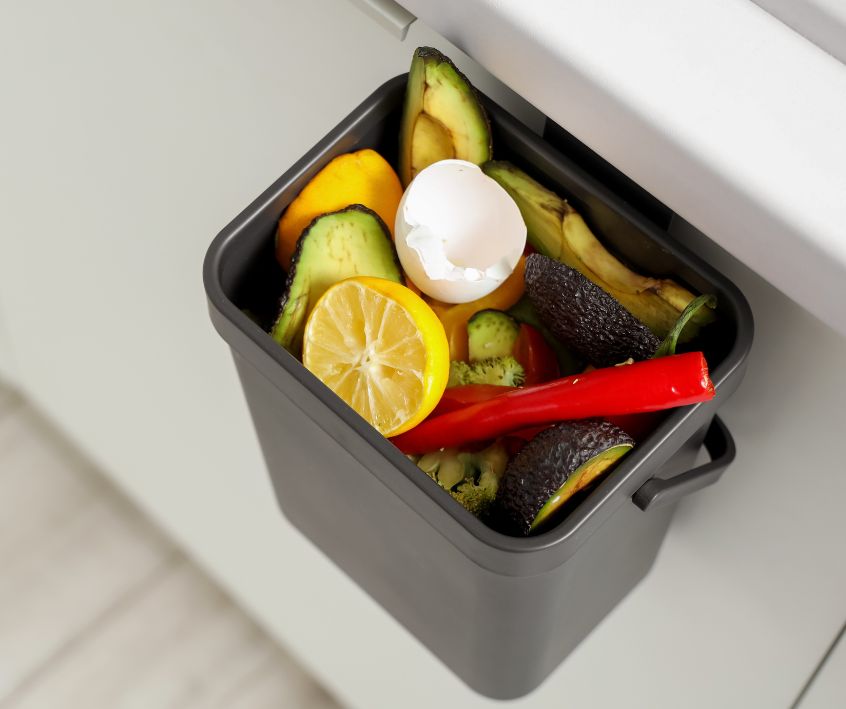A black compost bin with vegetable scraps mounted under a kitchen counter.