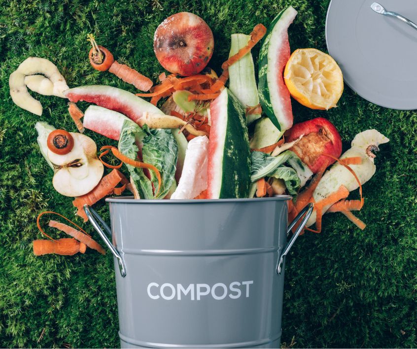 Peeled Vegetables and other kitchen scraps spilling out of a grey compost bin on green grass.