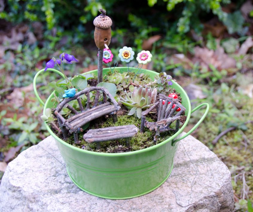 Fairy Garden Furniture consisting of a small wooden bench and table set in a small green metal bucket.