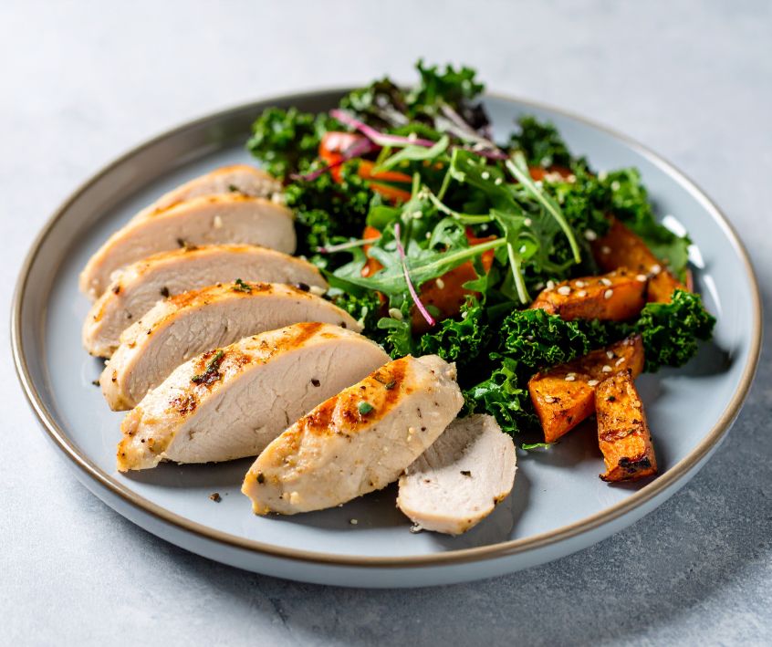 A plate of sliced grilled chicken breast with roasted sweet potatoes and warm kale salad on a white table.