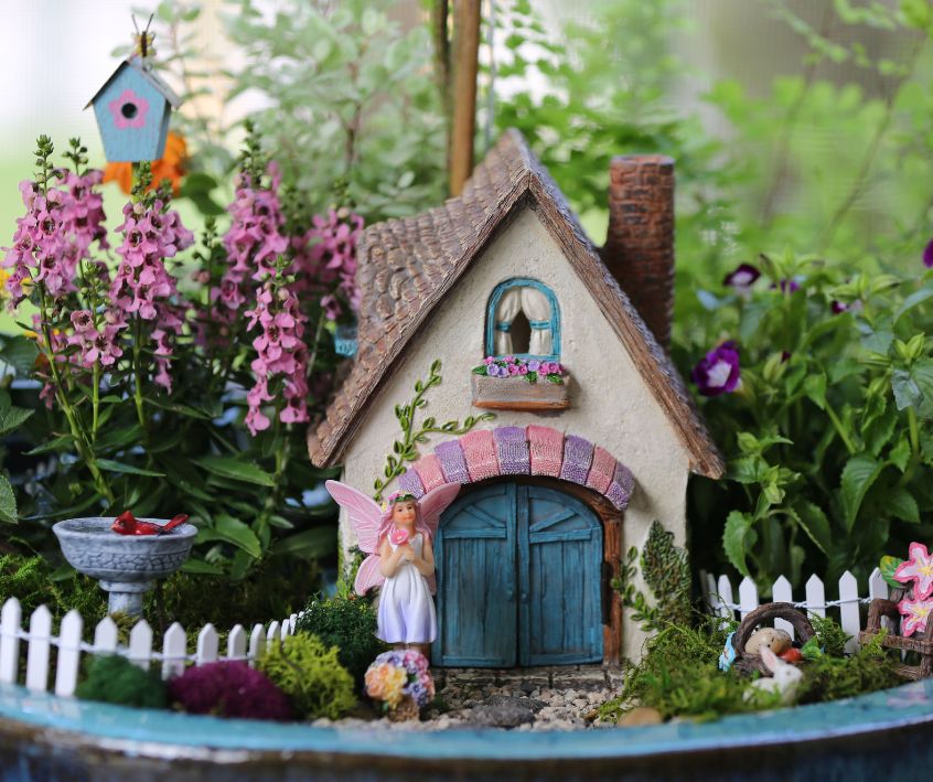 A miniature fairy garden with a fairy standing outside of a colorful home and bright flowers.