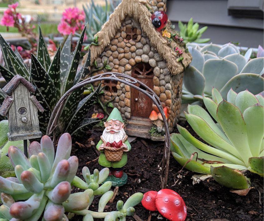 A small gnome in front of a stone home surrounds by lush succulents and an arch decorated with fairy lights.