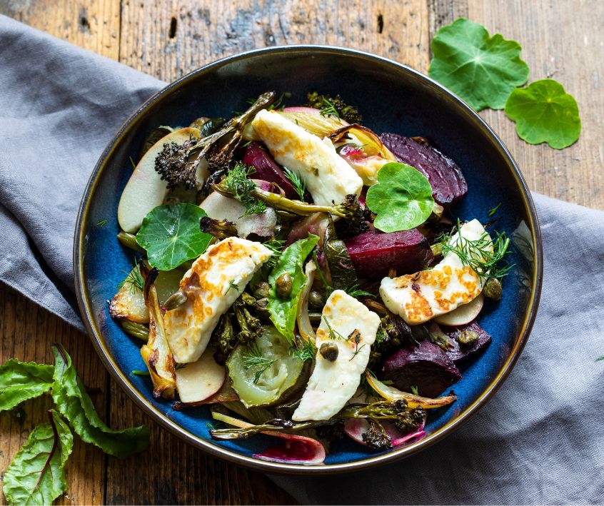 Warm salad with roast beetroot, broccoli, apple, fennel and crispy capers in a dark bowl on a alight blue fabric napkin.