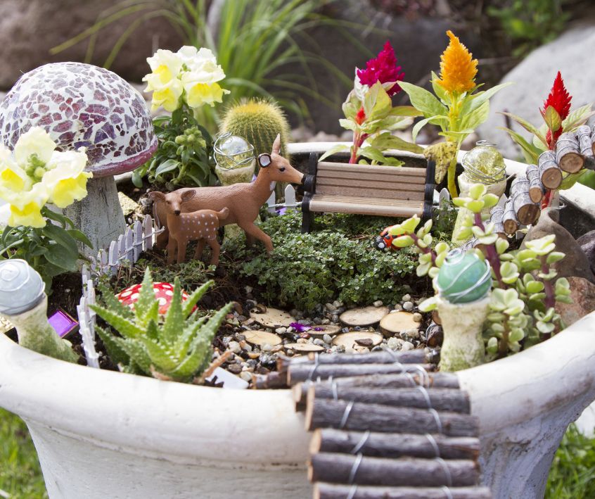 A fairy garden in a large pot featuring mini furniture and woodland creatures.